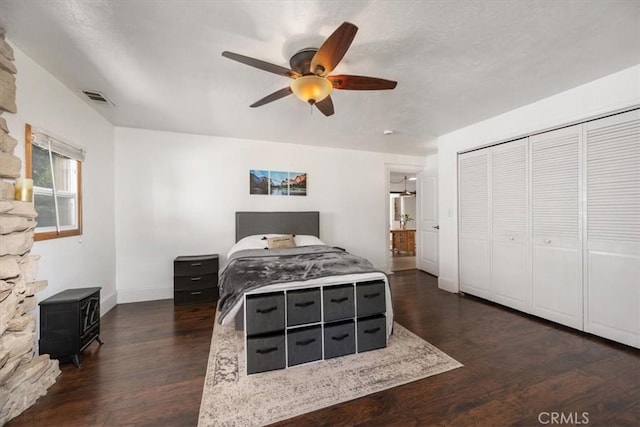 bedroom featuring ceiling fan, visible vents, a wood stove, dark wood finished floors, and baseboards