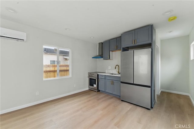 kitchen featuring light wood finished floors, a sink, a wall unit AC, wall chimney range hood, and appliances with stainless steel finishes