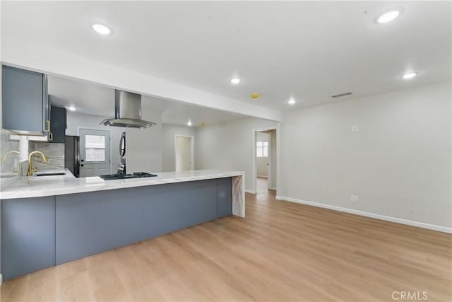 kitchen featuring backsplash, wall chimney range hood, a sink, light wood-type flooring, and a peninsula