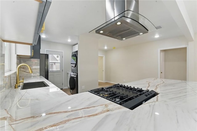 kitchen with light stone counters, a sink, island range hood, black appliances, and stacked washer and dryer