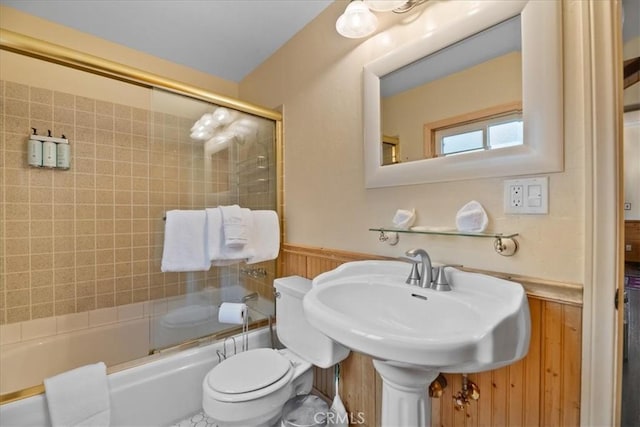 bathroom with a wainscoted wall, a sink, shower / bath combination with glass door, toilet, and wood walls