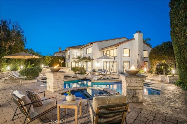 back of house at night with a chimney, a balcony, stucco siding, a patio area, and a pool with connected hot tub