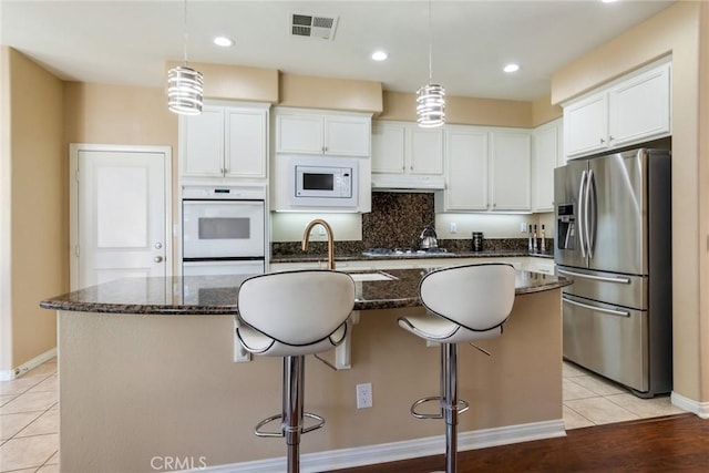 kitchen featuring white cabinets, dark stone countertops, a center island with sink, visible vents, and white appliances