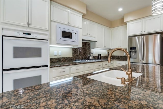 kitchen with a sink, dark stone countertops, white cabinetry, decorative backsplash, and white appliances