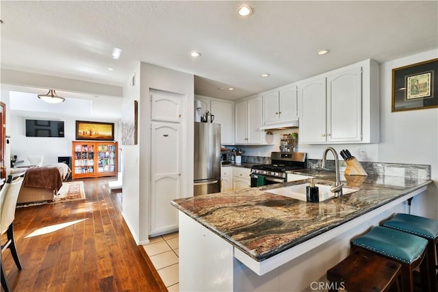 kitchen featuring appliances with stainless steel finishes, a sink, dark stone counters, a peninsula, and under cabinet range hood