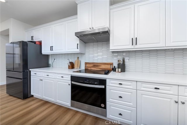 kitchen featuring wall oven, light wood-style flooring, black electric cooktop, under cabinet range hood, and freestanding refrigerator