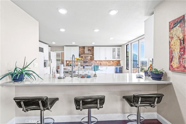 kitchen with wall chimney range hood, beverage cooler, a peninsula, and white cabinets