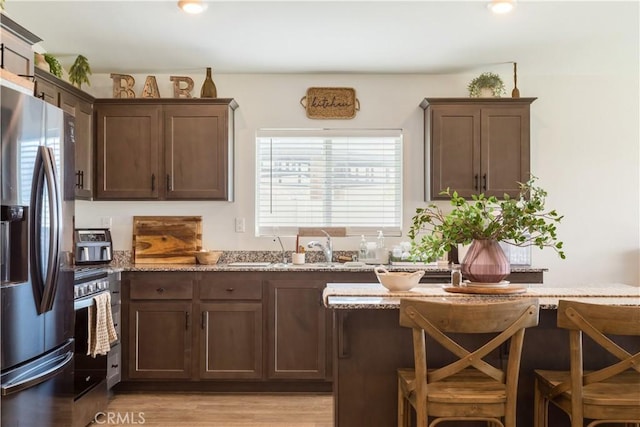 kitchen with light wood finished floors, appliances with stainless steel finishes, a breakfast bar, light stone counters, and dark brown cabinets