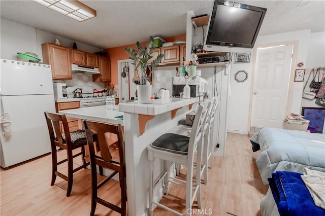 kitchen featuring tile counters, light wood finished floors, a kitchen breakfast bar, under cabinet range hood, and white appliances