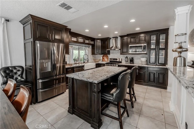 kitchen with dark brown cabinets, wall chimney range hood, stainless steel appliances, and visible vents