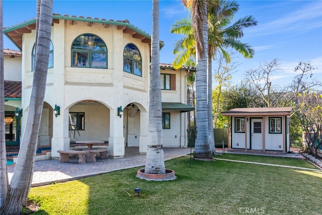 back of house featuring a tile roof, a patio area, a lawn, an outdoor structure, and stucco siding
