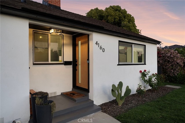 exterior entry at dusk featuring a chimney, roof with shingles, and stucco siding