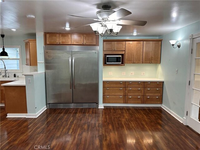 kitchen with built in appliances, brown cabinetry, dark wood-type flooring, a ceiling fan, and light countertops