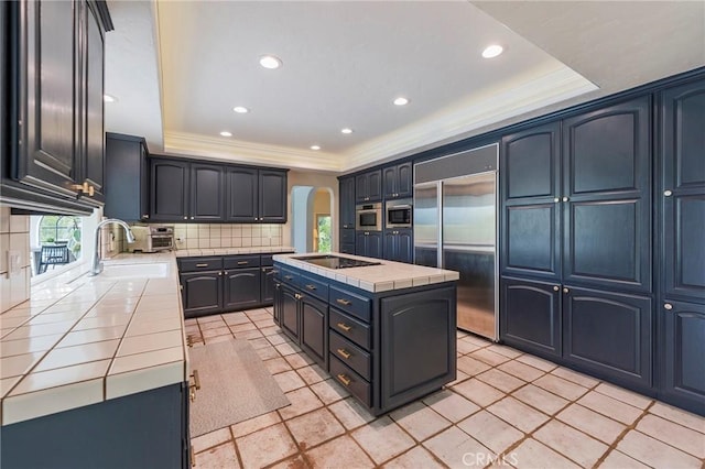 kitchen with a kitchen island with sink, tile countertops, crown molding, arched walkways, and built in appliances
