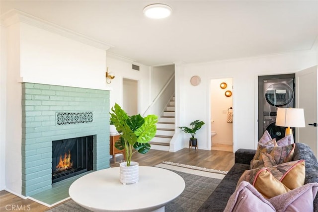 living room with stacked washer and dryer, a brick fireplace, wood finished floors, stairs, and ornamental molding