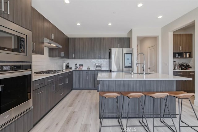 kitchen featuring a kitchen breakfast bar, under cabinet range hood, stainless steel appliances, light countertops, and a sink