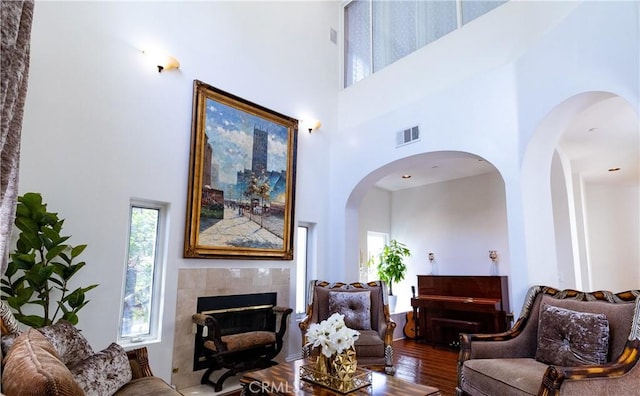 living area with visible vents, a towering ceiling, arched walkways, a tile fireplace, and wood finished floors