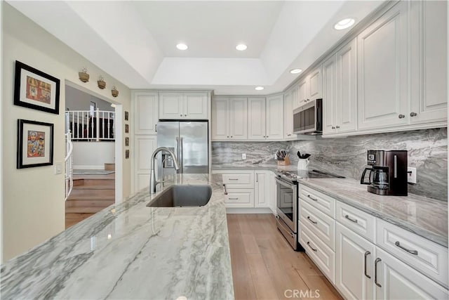 kitchen featuring a sink, light wood-type flooring, a raised ceiling, backsplash, and appliances with stainless steel finishes