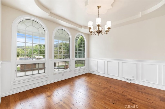 unfurnished dining area with a chandelier, a raised ceiling, wood-type flooring, and a wainscoted wall