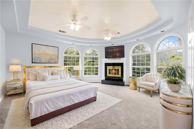 carpeted bedroom with a ceiling fan, a glass covered fireplace, a tray ceiling, and visible vents
