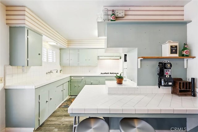 kitchen with tile counters, wood finished floors, a sink, backsplash, and under cabinet range hood