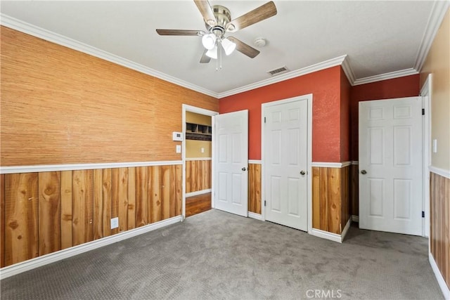 unfurnished bedroom featuring a wainscoted wall, carpet floors, wooden walls, crown molding, and ceiling fan