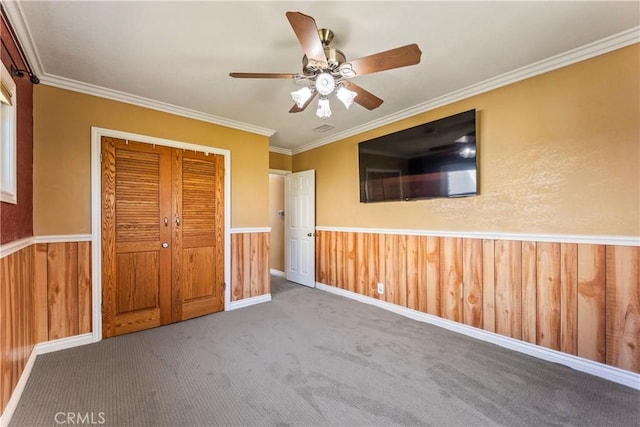 unfurnished bedroom featuring a wainscoted wall, a closet, carpet flooring, crown molding, and a ceiling fan