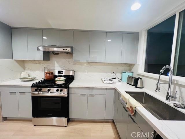 kitchen with decorative backsplash, a sink, gray cabinetry, under cabinet range hood, and stainless steel range with gas stovetop