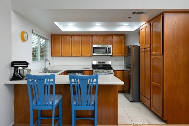 kitchen featuring appliances with stainless steel finishes, a raised ceiling, a peninsula, a sink, and brown cabinetry