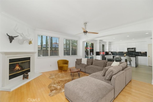 living room featuring ornamental molding, a glass covered fireplace, ceiling fan, light wood-type flooring, and recessed lighting
