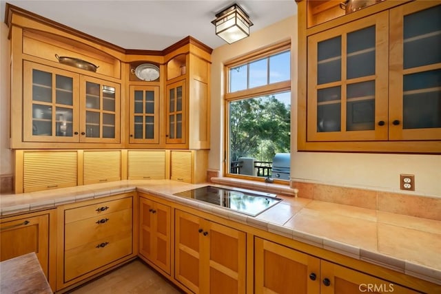 kitchen with tile counters, glass insert cabinets, and black electric stovetop