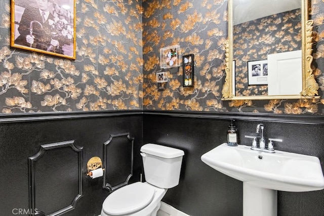 half bathroom with wallpapered walls, a wainscoted wall, a sink, and toilet
