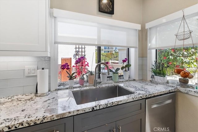 kitchen featuring light stone counters, a sink, decorative backsplash, dishwasher, and gray cabinetry