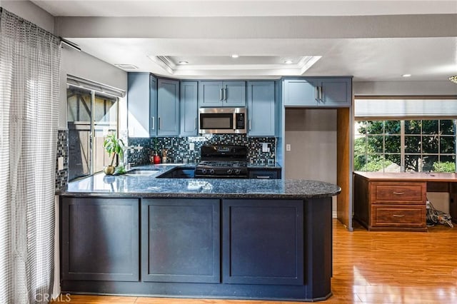 kitchen with dark stone counters, black range with gas stovetop, a raised ceiling, stainless steel microwave, and a peninsula