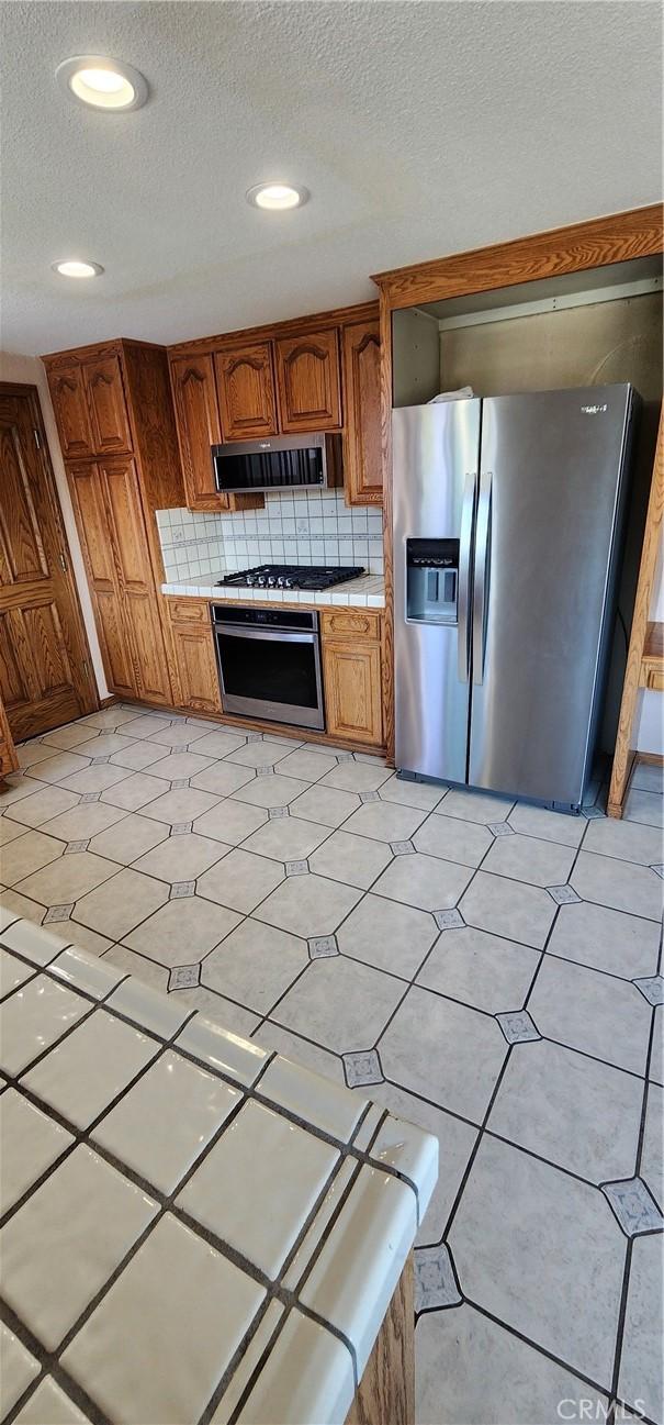 kitchen with brown cabinetry, decorative backsplash, tile countertops, a textured ceiling, and appliances with stainless steel finishes