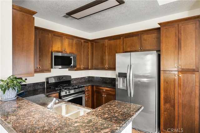 kitchen featuring a textured ceiling, a sink, dark stone countertops, a peninsula, and stainless steel appliances