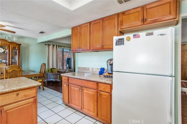 kitchen with brown cabinetry, a textured ceiling, visible vents, light tile patterned floors, and freestanding refrigerator