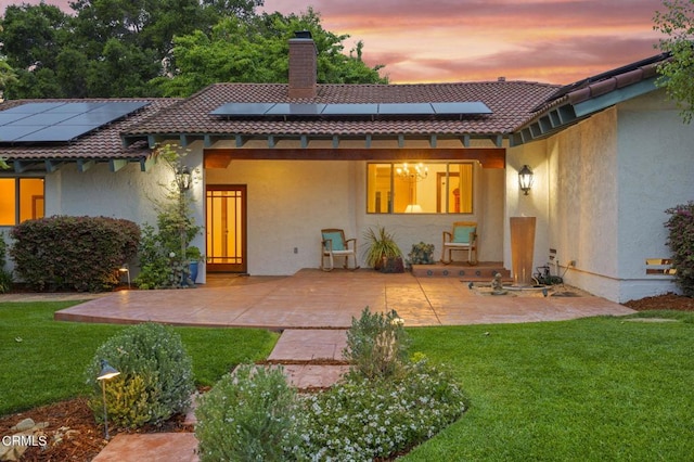 back of house at dusk featuring a patio, a lawn, a tiled roof, a chimney, and roof mounted solar panels
