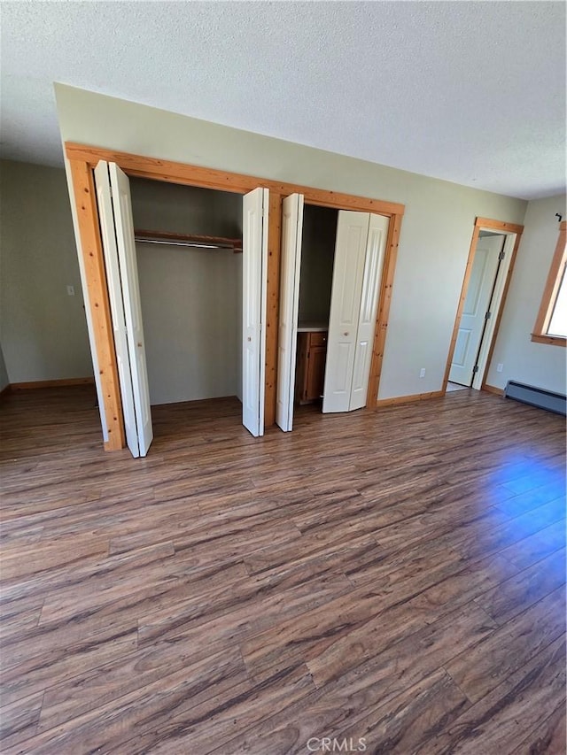 unfurnished bedroom with a textured ceiling, two closets, baseboards, wood finished floors, and a baseboard heating unit