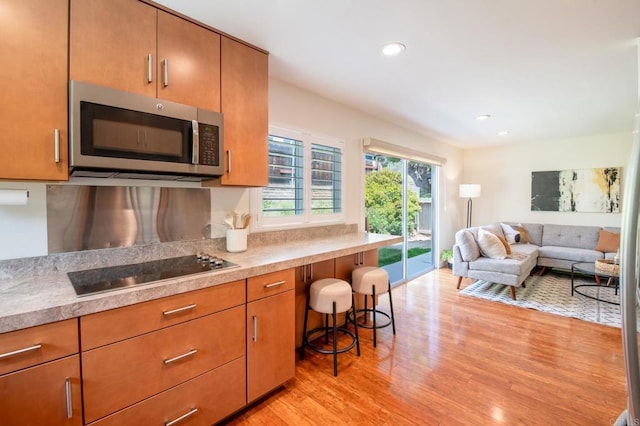 kitchen with stainless steel microwave, black electric cooktop, light wood-type flooring, brown cabinetry, and open floor plan