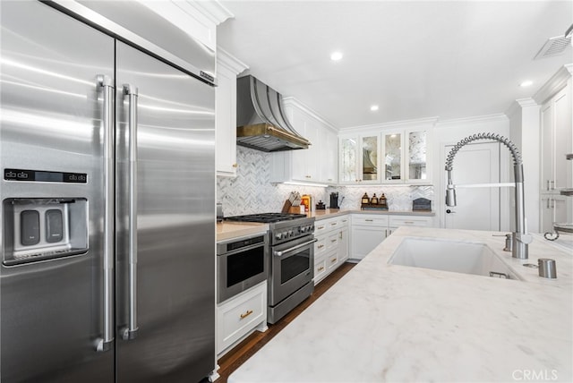 kitchen featuring wall chimney exhaust hood, a sink, premium appliances, decorative backsplash, and light stone counters