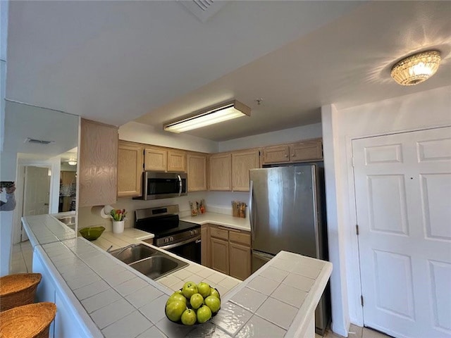 kitchen with light brown cabinets, stainless steel appliances, tile counters, a sink, and a peninsula