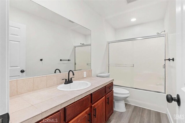 bathroom with recessed lighting, wood finished floors, combined bath / shower with glass door, toilet, and vanity