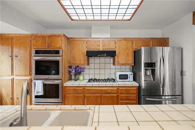 2116 Ironwood Ct, Pine Mountain Club, CA 93222 - kitchen with brown cabinets, under cabinet range hood, stainless steel appliances, backsplash, and tile countertops