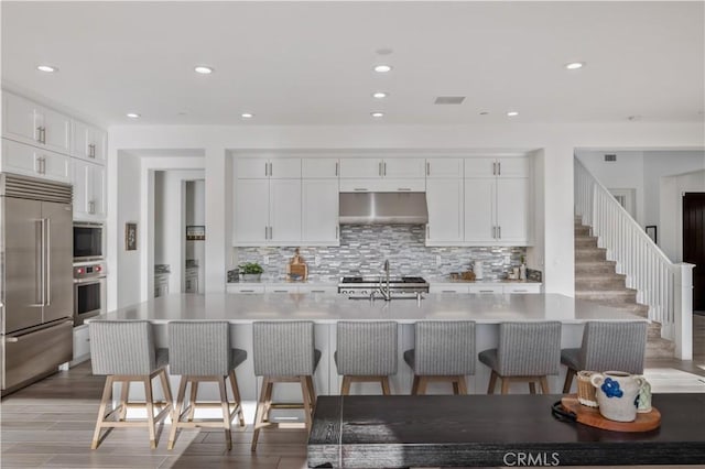 kitchen with backsplash, a sink, light countertops, built in appliances, and under cabinet range hood