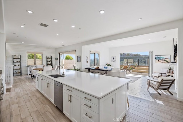 kitchen with stainless steel dishwasher, a center island with sink, wood finish floors, a sink, and white cabinetry