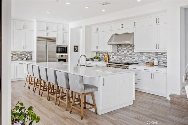 kitchen with decorative backsplash, built in appliances, light wood-style floors, under cabinet range hood, and a sink