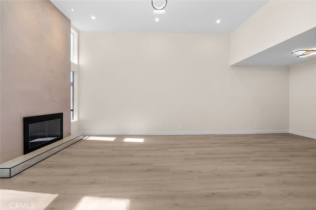 unfurnished living room featuring recessed lighting, light wood-style flooring, baseboard heating, a glass covered fireplace, and baseboards