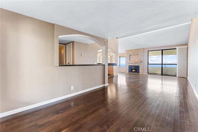 unfurnished living room with baseboards, wood finished floors, arched walkways, a stone fireplace, and ornate columns
