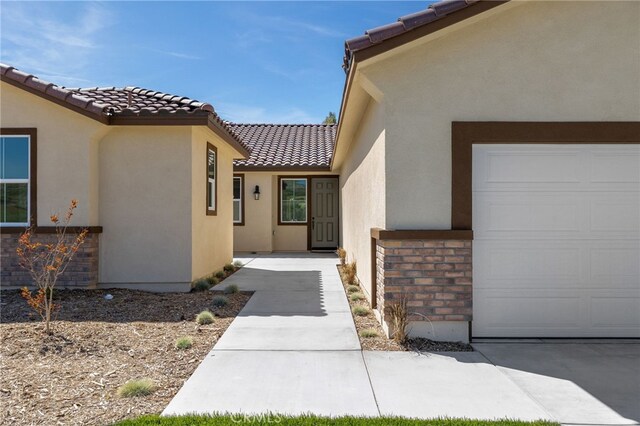 doorway to property featuring an attached garage, a tiled roof, brick siding, and stucco siding